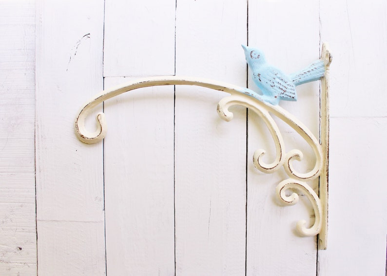 May include: White metal wall-mounted plant hanger with a decorative scroll design and a light blue bird figurine.