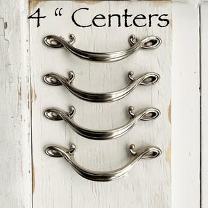 May include: Four silver cabinet pulls with a decorative scroll design. The pulls are mounted on a white wooden board with the text "4" Centers" written in black.