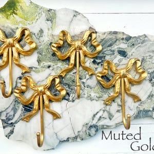 May include: Four gold-colored bow-shaped wall hooks. The hooks are mounted on a decorative stone slab with green and white marbling. The text "Muted Gold" is visible in the lower right corner.
