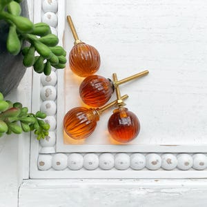 May include: Four round, amber-colored glass cabinet knobs with vertical ribbed textures. Each knob features a gold-toned metal base and a threaded screw post extending from the back, designed for attachment to furniture or cabinetry doors.