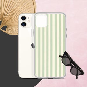 May include: A white iPhone with a clear case featuring vertical light green and white stripes. Black cat-eye sunglasses rest on a pink and black background. A dried palm leaf fan is in the background.