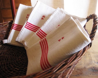 May include: A set of four white linen tea towels with red stripes and the letter "A" embroidered in red. The towels are folded and placed in a wicker basket.