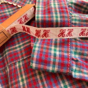 May include: A close-up of a red, white, and blue plaid fabric with a white ribbon sewn on it. The ribbon has the text "H.H." repeated on it.
