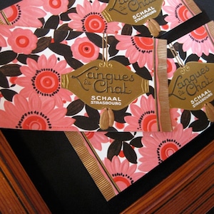 May include: Two vintage packaging labels for "Langues de Chat" cookies. The labels feature a black and white floral design with pink flowers and gold accents. The text "Langues de Chat" is written in a decorative script font and the company name "Schaal Strasbourg" is printed below.