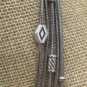 May include: Silver-toned necklace with multiple strands. The necklace features a geometric diamond-shaped pendant and cylindrical beads. The strands are made of a snake chain design. The necklace is displayed against a textured, neutral background.
