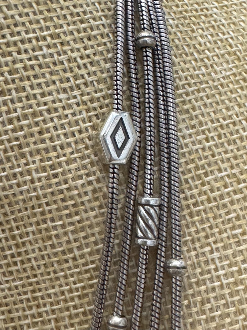 May include: Silver-toned necklace with multiple strands. The necklace features a geometric diamond-shaped pendant and cylindrical beads. The strands are made of a snake chain design. The necklace is displayed against a textured, neutral background.
