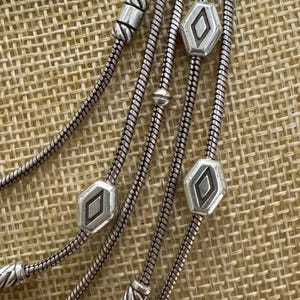 May include: Close-up of a silver-toned necklace with multiple strands. The necklace features decorative geometric beads and a snake chain design. The beads have a diamond-shaped design in the center.