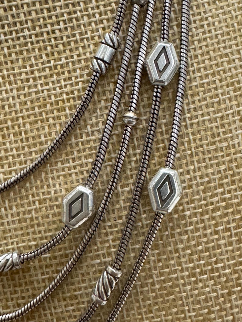 May include: Close-up of a silver-toned necklace with multiple strands. The necklace features decorative geometric beads and a snake chain design. The beads have a diamond-shaped design in the center.