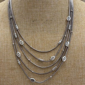 May include: A multi-strand silver necklace with a layered design. The necklace features five strands of varying lengths, adorned with diamond-shaped and round beads. The necklace is displayed on a neutral-toned mannequin.