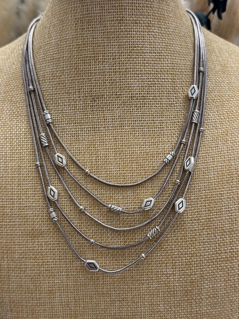 May include: A multi-strand silver necklace with a layered design. The necklace features five strands of varying lengths, adorned with diamond-shaped and round beads. The necklace is displayed on a neutral-toned mannequin.