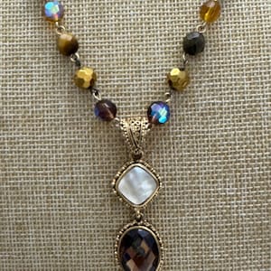 Pode incluir: Um colar dourado com um pendente com uma pedra quadrada de madrepérola sobre uma gema oval castanha. O colar tem uma corrente de contas de várias cores, incluindo contas douradas, castanhas e iridescentes. O colar é exibido numa superfície texturizada e bege.