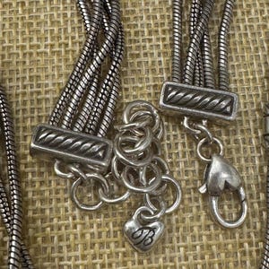 May include: Close-up of a silver-toned necklace with multiple strands. The necklace features decorative clasps and a heart-shaped charm with the letter "B". The background is a textured, neutral fabric.