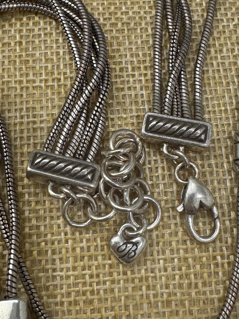 May include: Close-up of a silver-toned necklace with multiple strands. The necklace features decorative clasps and a heart-shaped charm with the letter "B". The background is a textured, neutral fabric.