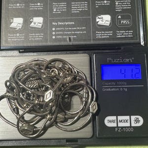May include: A silver necklace with geometric charms is displayed on a digital scale. The scale reads 41.2, with a capacity of 1000g and a graduation of 0.1g. The scale's buttons include TARE, MODE, and ON/OFF.
