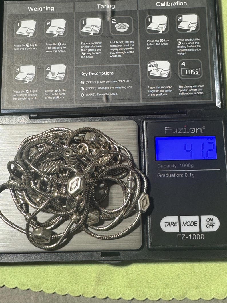 May include: A silver necklace with geometric charms is displayed on a digital scale. The scale reads 41.2, with a capacity of 1000g and a graduation of 0.1g. The scale's buttons include TARE, MODE, and ON/OFF.