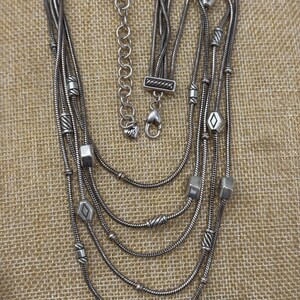 May include: Silver-toned multi-strand necklace with geometric and beaded accents. The necklace features a variety of shapes, including cubes, diamonds, and cylindrical beads. The necklace has a lobster clasp closure and an adjustable chain.