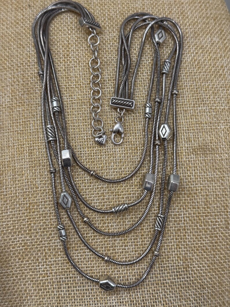 May include: Silver-toned multi-strand necklace with geometric and beaded accents. The necklace features a variety of shapes, including cubes, diamonds, and cylindrical beads. The necklace has a lobster clasp closure and an adjustable chain.