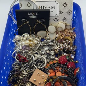 Op de afbeelding: Een blauwe plastic mand vol met diverse sieraden. De collectie omvat oorbellen, kettingen, armbanden en broches in zilver, goud en gemengde metaaltinten. Sommige stukken hebben parels, strass en kleurrijke accenten. Een doos met de tekst "SHIVAM" is ook zichtbaar.