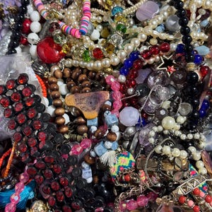 Puede incluir: Primer plano de una colección de collares y pulseras. La joyería incluye hilos de perlas, cuentas y acentos coloridos. Hay cuentas rojas, negras, blancas y azules. Algunas piezas tienen detalles de estrellas y borlas. La joyería está en una cesta morada.