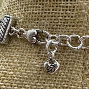 May include: Close-up of a silver-toned necklace with a heart-shaped charm. The charm has a cursive "B" on it. The necklace has a textured clasp and a series of oval links. The background is a tan woven fabric.
