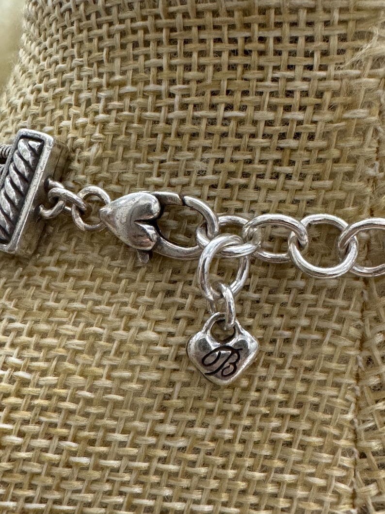 May include: Close-up of a silver-toned necklace with a heart-shaped charm. The charm has a cursive "B" on it. The necklace has a textured clasp and a series of oval links. The background is a tan woven fabric.