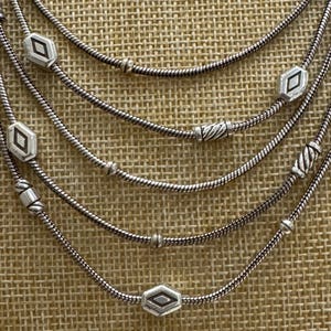 May include: A multi-strand silver necklace with geometric diamond-shaped and cylindrical beads. The necklace has a delicate, layered design, with each strand featuring a different arrangement of beads. The necklace is displayed against a textured, neutral-toned background.