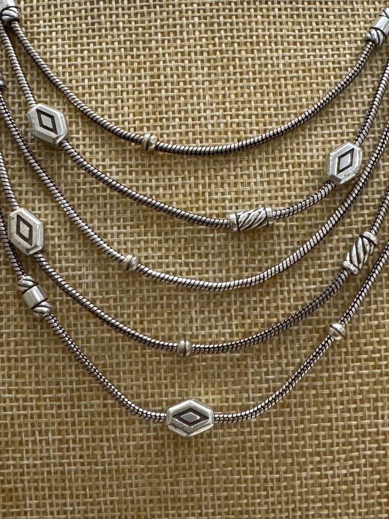 May include: A multi-strand silver necklace with geometric diamond-shaped and cylindrical beads. The necklace has a delicate, layered design, with each strand featuring a different arrangement of beads. The necklace is displayed against a textured, neutral-toned background.