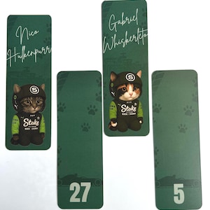 May include: Green rectangular bookmarks featuring cartoon cats in racing attire. One bookmark has the name "Nico Hulkenpurr", another "Gabriel Whiskerto". The other two bookmarks have the numbers 27 and 5. The bookmarks have paw prints.