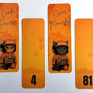 May include: Orange and yellow bookmarks featuring cartoon cats in racing suits and helmets. The bookmarks have the names "Lando Pawris" and "Oscar Meowstry" written on them, along with the numbers "4" and "81".