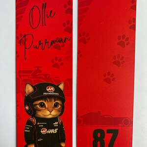 May include: Red bookmark featuring a cartoon cat in a racing suit and helmet. The cat is wearing a black and red racing suit with sponsor logos. The bookmark also has the name "Ollie Purrman" and the number "87".