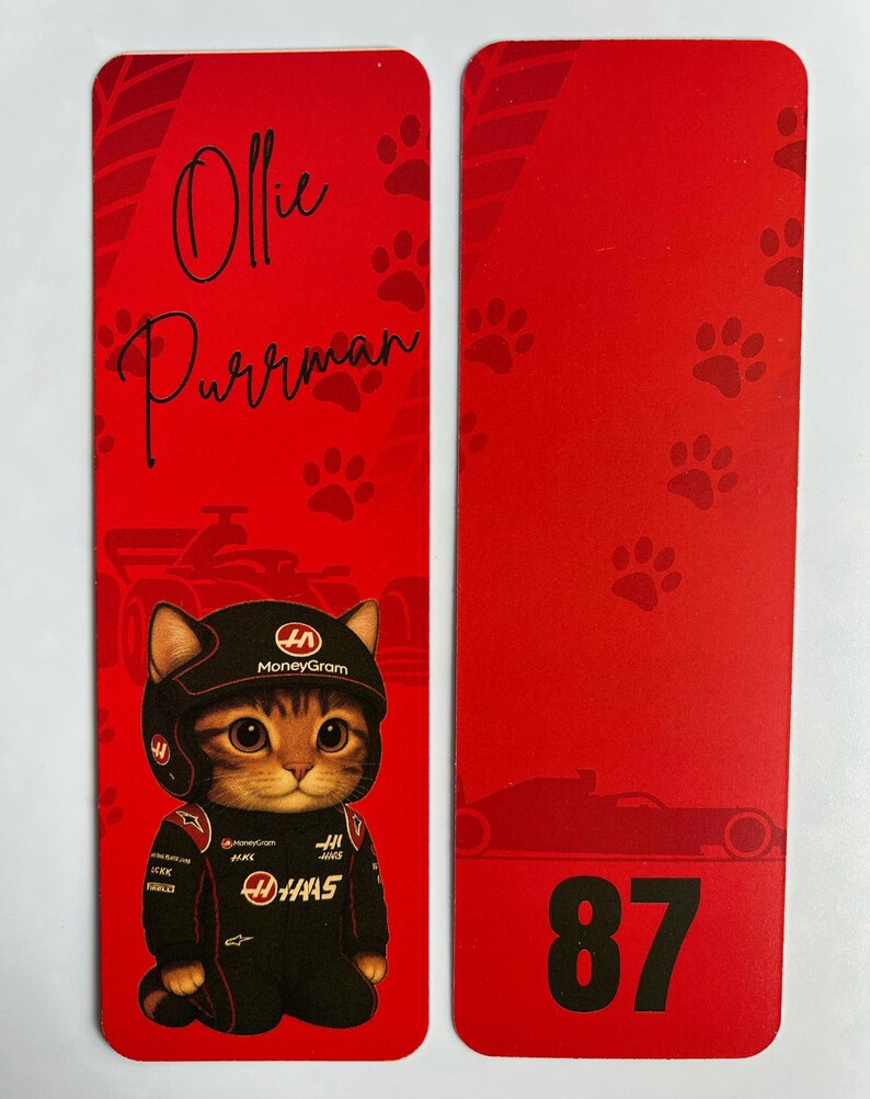 May include: Red bookmark featuring a cartoon cat in a racing suit and helmet. The cat is wearing a black and red racing suit with sponsor logos. The bookmark also has the name "Ollie Purrman" and the number "87".