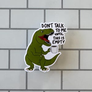 May include: A sticker featuring a green cartoon dinosaur holding a coffee mug. The dinosaur has an open mouth with red inside. Text on the sticker reads "DON'T TALK TO ME UNTIL THIS IS EMPTY."
