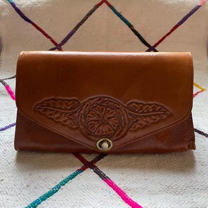 May include: A brown leather clutch with a floral design embossed on the flap. The clutch has a rectangular shape with a rounded top and a metal clasp closure. The leather has a glossy finish, and the background is a patterned textile.