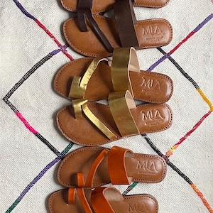 Pode incluir: Uma coleção de sandálias de couro em várias cores, incluindo verde-oliva, castanho, dourado e laranja. Cada sandália apresenta sola e tiras de couro, com a marca "MIA" em relevo na palmilha. As sandálias estão dispostas sobre um tapete estampado.