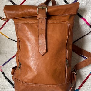 May include: A brown leather backpack with a roll-top closure and a buckle. The bag has two zippered side pockets and adjustable shoulder straps. The leather has a textured appearance. The backpack is sitting on a patterned rug.