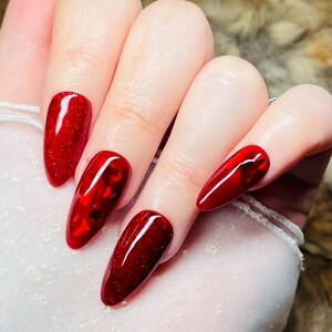 Puede incluir: Una mano con uñas postizas largas, rojas y brillantes. Las uñas tienen forma puntiaguda y un acabado brillante. Algunas uñas tienen un efecto brillante, mientras que otras tienen detalles en forma de corazón.
