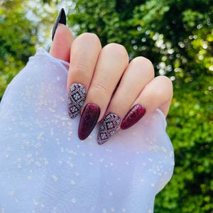 May include: A hand displaying long, almond-shaped artificial nails. The nails are painted in a deep burgundy color, with two accent nails featuring a white floral pattern. One nail is painted black.