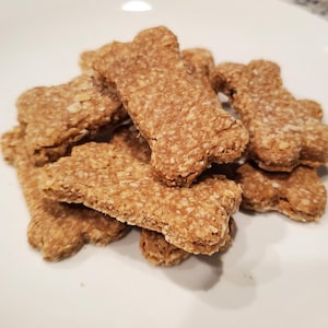May include: A pile of bone-shaped dog biscuits. The biscuits are a light brown color and appear to be made with oats or a similar grain. They are stacked on a white plate, suggesting a homemade or artisanal treat.