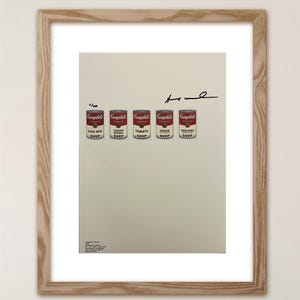 Puede incluir: Una impresión enmarcada que muestra cinco latas de sopa Campbell's en rojo y blanco, etiquetadas como Black Bean, Chicken Noodle, Tomato, Onion y Vegetable. La impresión incluye una firma, el número de edición 0/100 y texto pequeño en un marco de madera clara.