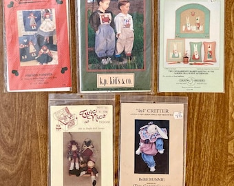 May include: Five vintage craft pattern packets in plastic sleeves. The packets feature images of dolls, children, and rabbits. Titles include "Country Stitches", "k.p. kid's & co.", "Rabbits in the Garden", "Twice as Nice Designs", and "Bebe Bunnie".