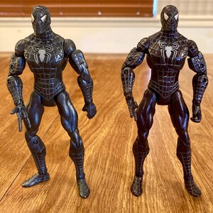 May include: Two black Spider-Man action figures. The figures are in a standing pose, with detailed textures and the iconic spider emblem on the chest. The figures are on a wooden surface.