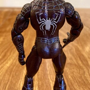 May include: A brown action figure of a superhero, viewed from the back. The figure has a textured, patterned suit with a white spider emblem on the back. The figure is standing on a wooden surface.