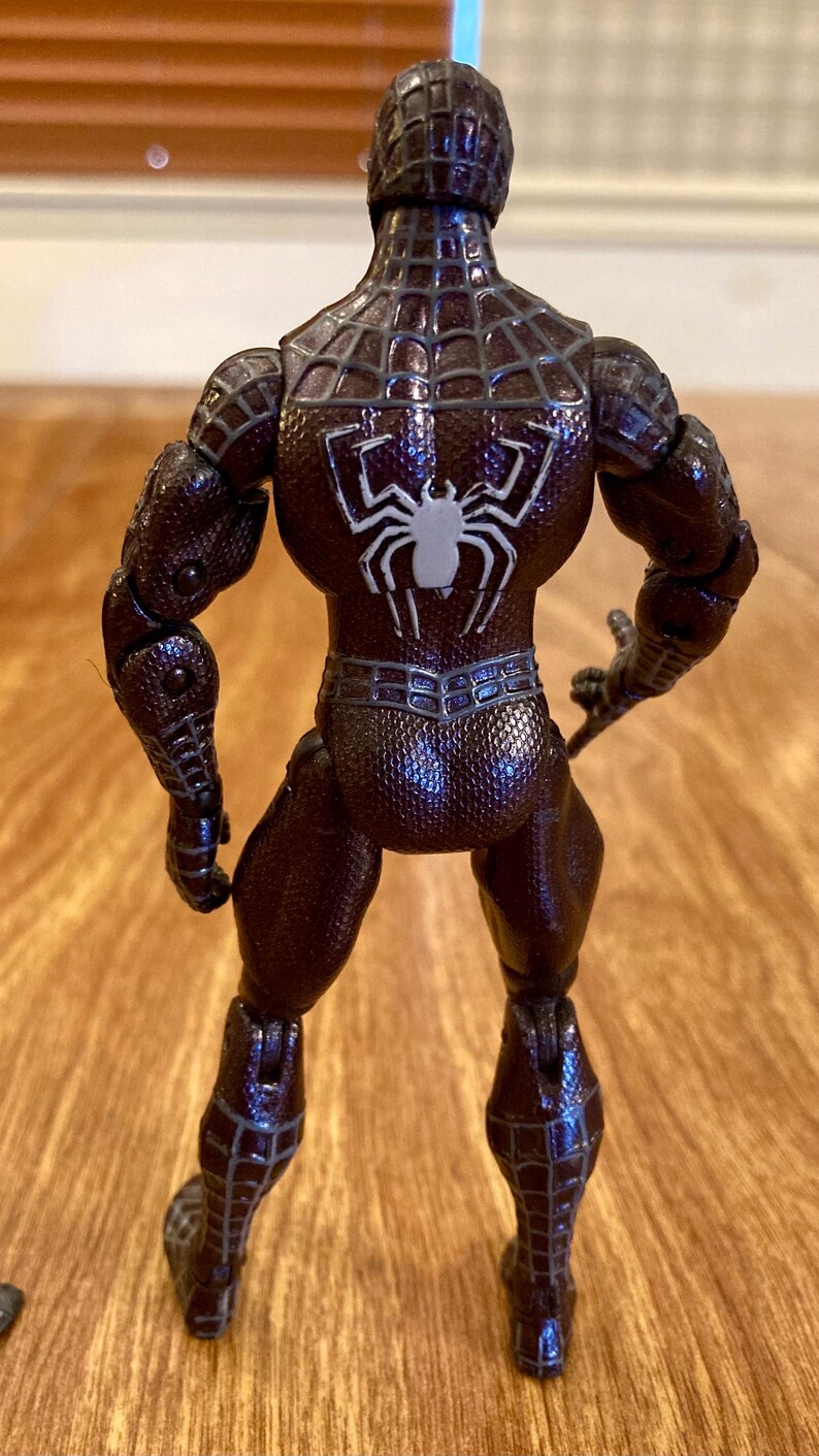 May include: A brown action figure of a superhero, viewed from the back. The figure has a textured, patterned suit with a white spider emblem on the back. The figure is standing on a wooden surface.