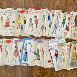 May include: A collection of vintage Simplicity sewing pattern envelopes, featuring illustrations of clothing designs. The patterns showcase various styles, including dresses, skirts, and tops, with numbers visible on the covers. The envelopes are arranged on a wooden surface.