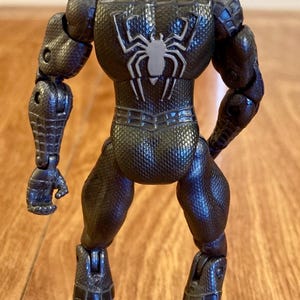 May include: A black action figure of a superhero, viewed from the back. The figure has a textured, web-like pattern and a white spider emblem on the back. The figure is articulated at the joints.