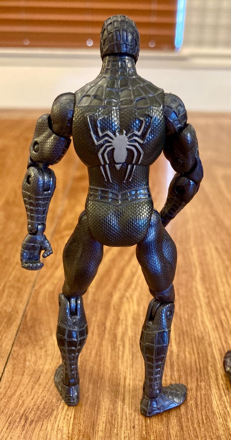 May include: A black action figure of a superhero, viewed from the back. The figure has a textured, web-like pattern and a white spider emblem on the back. The figure is articulated at the joints.
