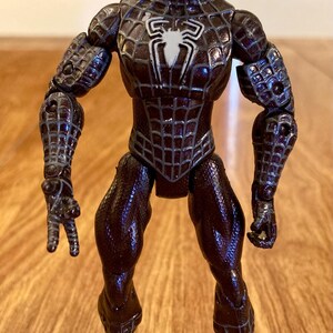 May include: A Spider-Man action figure in a dark brown and black color scheme. The figure has a spider web pattern and a white spider emblem on the chest. The figure is standing on a wooden surface.