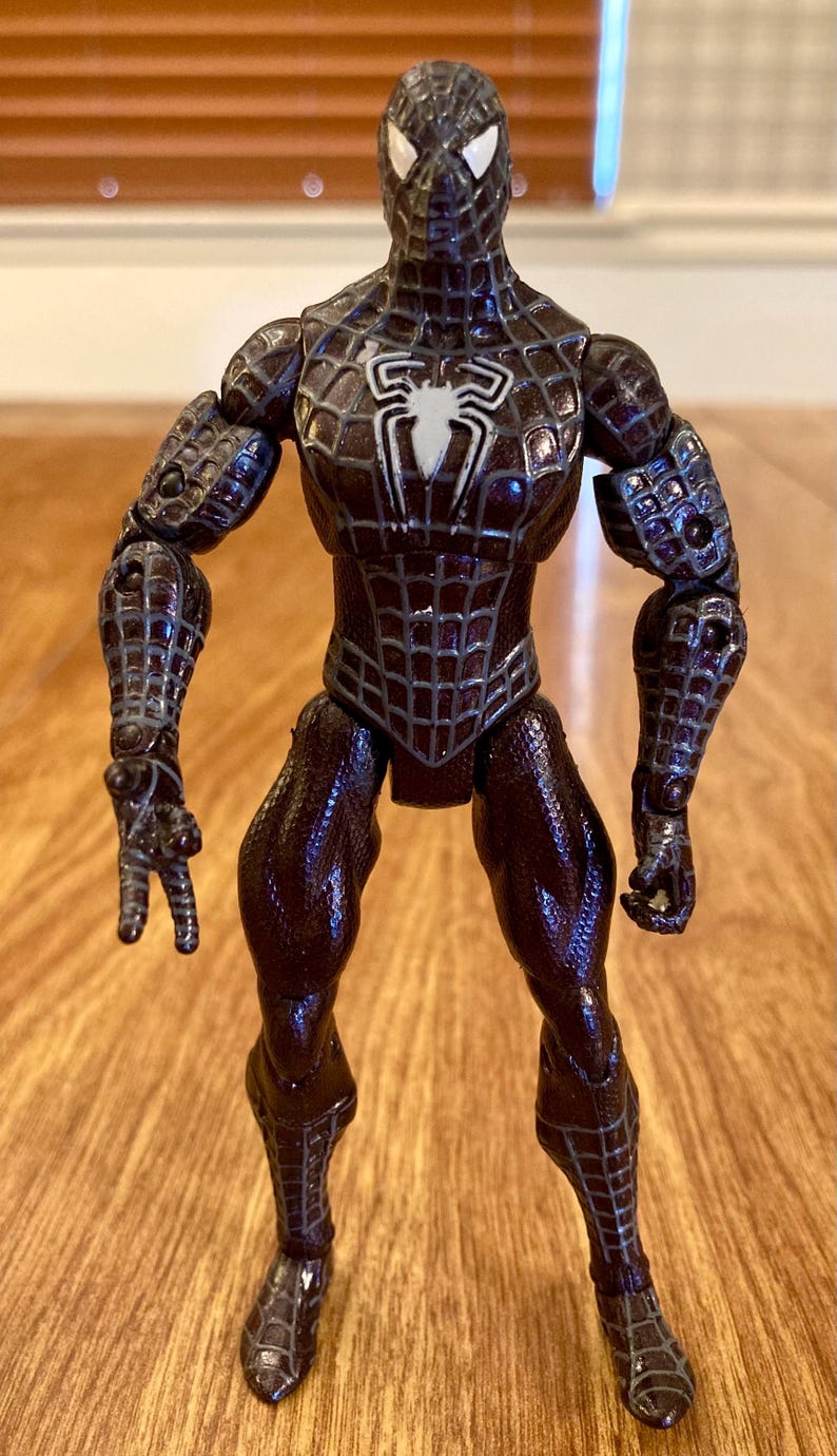 May include: A Spider-Man action figure in a dark brown and black color scheme. The figure has a spider web pattern and a white spider emblem on the chest. The figure is standing on a wooden surface.