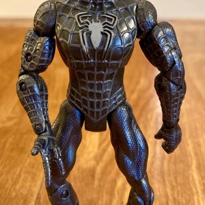 May include: A black Spider-Man action figure with a textured, web-like pattern. The figure has a white spider emblem on the chest and silver eye details. The figure is posed with arms out, showcasing the detailed design.