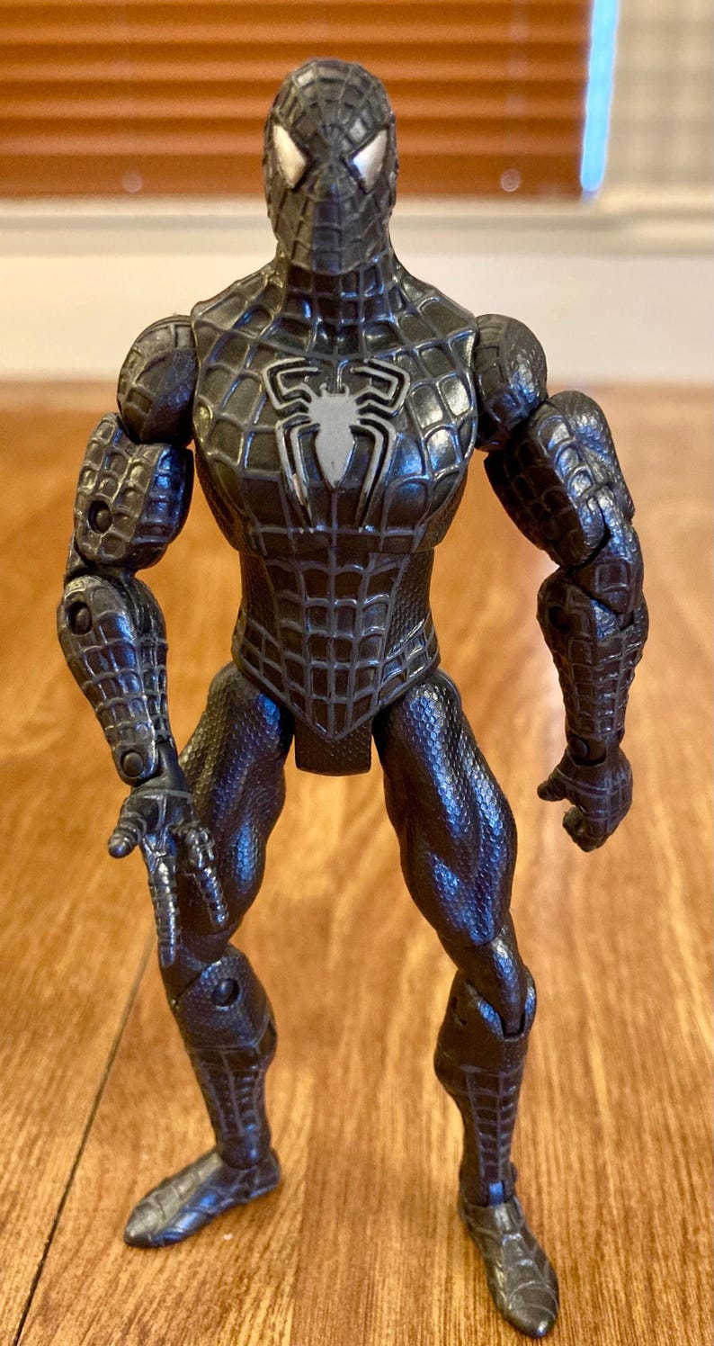 May include: A black Spider-Man action figure with a textured, web-like pattern. The figure has a white spider emblem on the chest and silver eye details. The figure is posed with arms out, showcasing the detailed design.